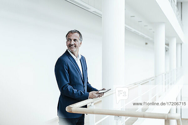 Lächelnder männlicher Geschäftsmann  der ein digitales Tablet an einem Geländer im Büroflur hält
