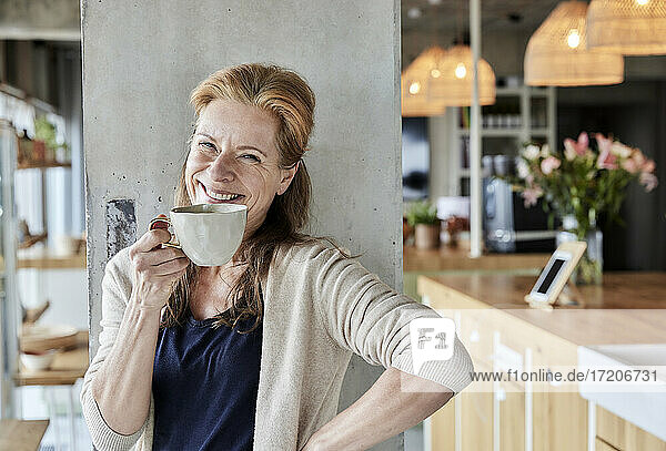 Happy mature woman with hand on hip having coffee against column at home
