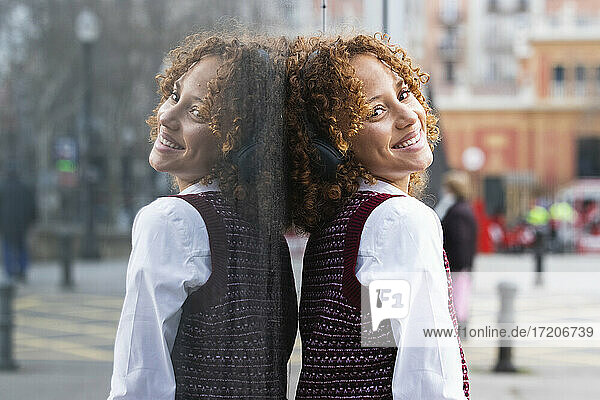 Lächelndes Teenager-Mädchen mit Kopfhörern  das sich an eine Glaswand lehnt