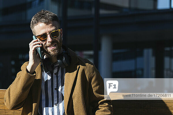 Geschäftsmann mit Sonnenbrille  der ein Mobiltelefon benutzt  während er auf einer Bank sitzt
