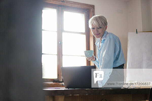 Lächelnde Geschäftsfrau  die eine Kaffeetasse hält  während sie einen Laptop auf dem Schreibtisch im Büro zu Hause benutzt