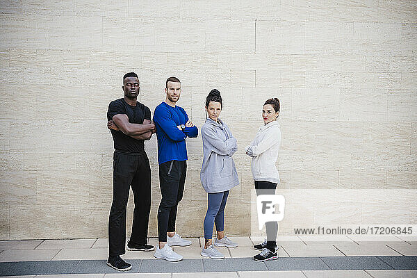 Selbstbewusste multiethnische Gruppe von Sportlern mit verschränkten Armen auf dem Fußweg an der Mauer stehend