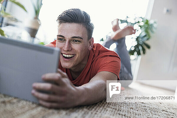 Fröhlicher Mann  der auf dem Teppich liegt  während er im Wohnzimmer zu Hause Videos ansieht