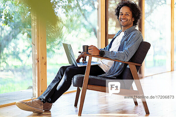 Junger Mann mit Laptop und Kaffeetasse  der lächelnd im Vorgarten sitzt
