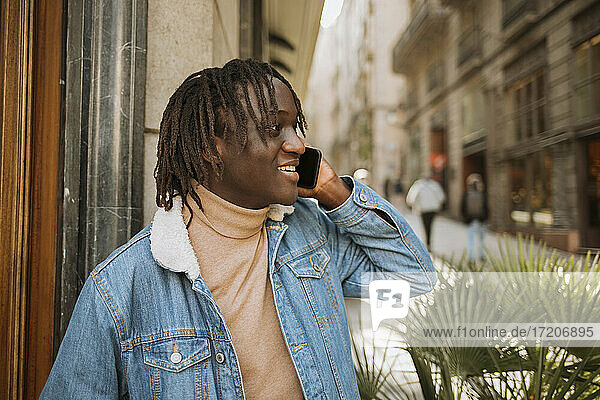 Cheerful young man with locs hairstyle talking on phone while looking away in city
