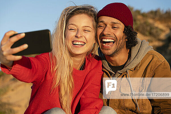 Glückliches Paar lächelnd während der Aufnahme von Selfie durch Handy sitzen im Freien