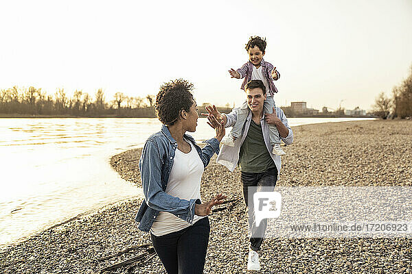 Young woman gesturing to man running with boy on shoulder by lake