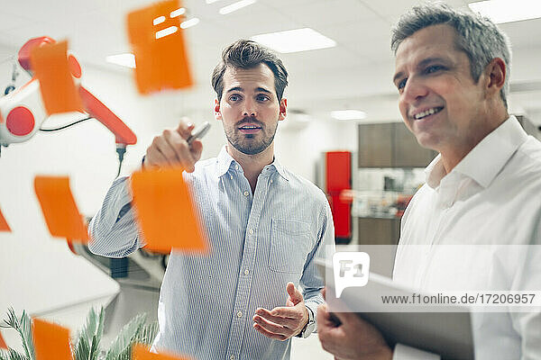 Young male engineer explaining business plans to smiling mature man in front of adhesive notes on glass wall in factory