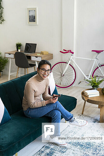 Smiling woman with mobile phone sitting on sofa at home