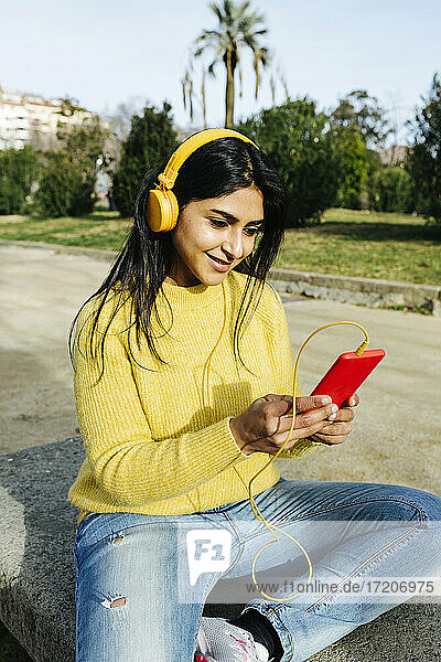 Lächelnde Frau  die ein Mobiltelefon benutzt  während sie in einem öffentlichen Park über Kopfhörer Musik hört