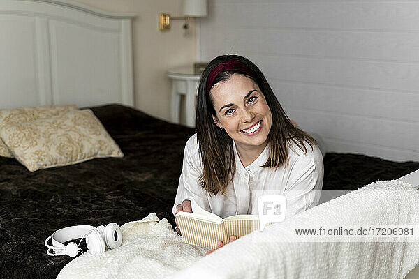 Lächelnde Frau mit Buch auf dem Bett liegend zu Hause