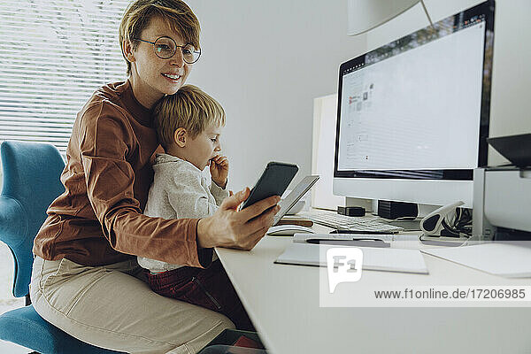 Mutter benutzt ihr Smartphone  während ihr Sohn im Home Office auf dem Schoß sitzt