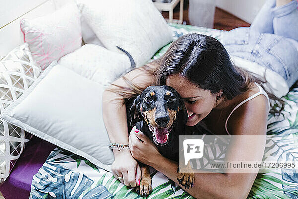 Smiling woman embracing Dachshund dog while lying down on bed at home