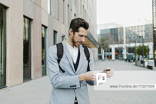 Male entrepreneur with backpack using mobile phone while standing against building