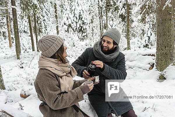 Young couple smiling while having coffee in forest during winter