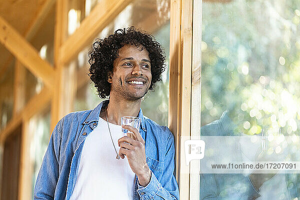 Lächelnder Mann mit Glas  der wegschaut und sich an ein Fenster im Vorgarten lehnt