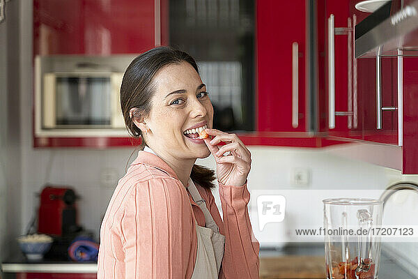 Cheerful woman eating slice of strawberry in kitchen
