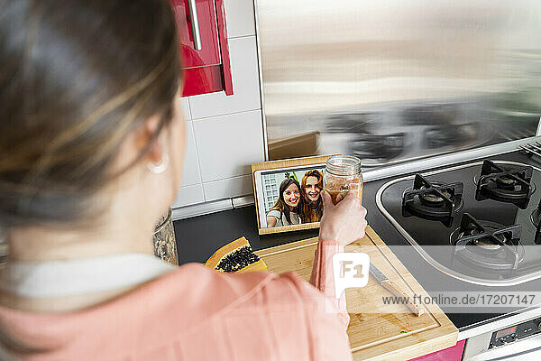 Young woman showing smoothie to friends on video call from kitchen