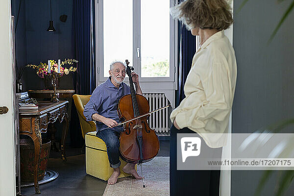 Lächelnder Mann schaut Frau an  während er zu Hause Cello spielt