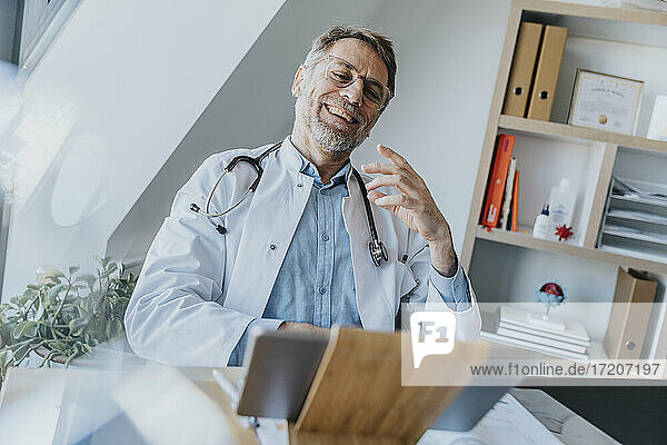 Männlicher Angestellter des Gesundheitswesens führt ein Videogespräch mit einem Patienten über ein digitales Tablet  während er in der Arztpraxis sitzt