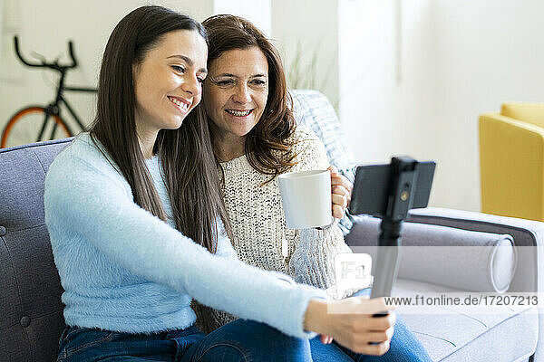 Smiling daughter and mother vlogging through smart phone while sitting on sofa at home