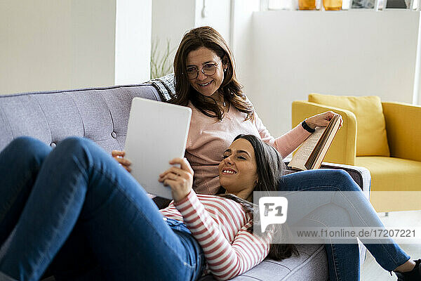 Lächelnde Tochter zeigt digitales Tablet  während sie auf dem Schoß der Mutter mit einem Buch auf dem Sofa zu Hause sitzt