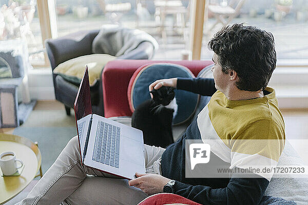 Mittlerer erwachsener Mann mit Laptop  der seine Katze streichelt  während er zu Hause auf dem Sofa sitzt