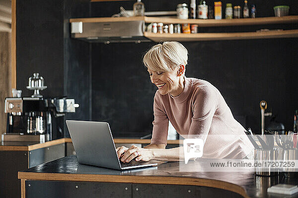 Happy businesswoman working on laptop at kitchen counter in home office