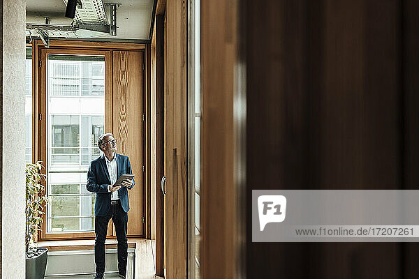 Älterer Geschäftsmann mit digitalem Tablet  der im Bürokorridor steht und nach oben schaut