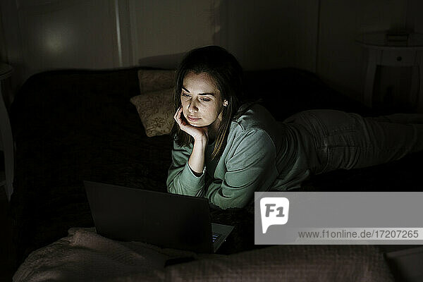 Junge Frau schaut auf einen Laptop  während sie zu Hause auf dem Bett liegt