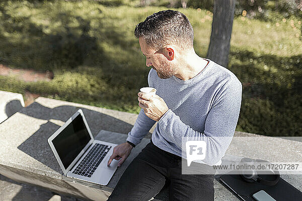 Älterer Geschäftsmann mit Einweg-Kaffeebecher und Laptop  während er auf einer Betonbank in einem öffentlichen Park sitzt