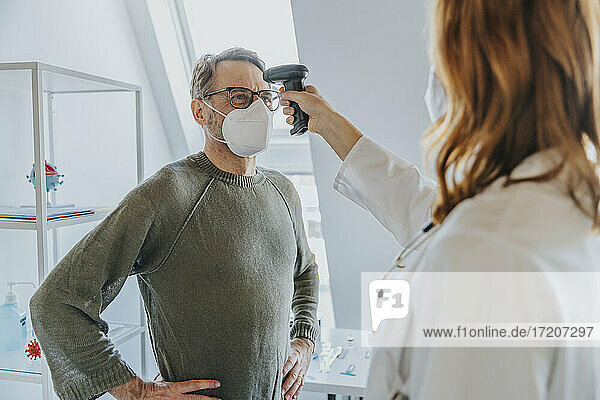 Ein Arzt überprüft einen Patienten  der mit der Hand auf der Taille steht  mit einem Infrarotthermometer im Untersuchungsraum