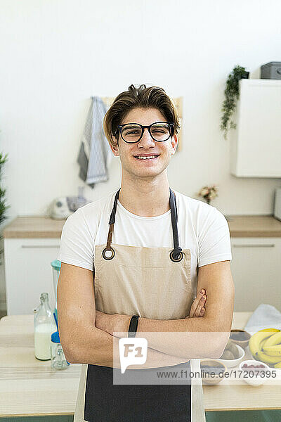 Handsome smiling man in apron standing with arms crossed in kitchen
