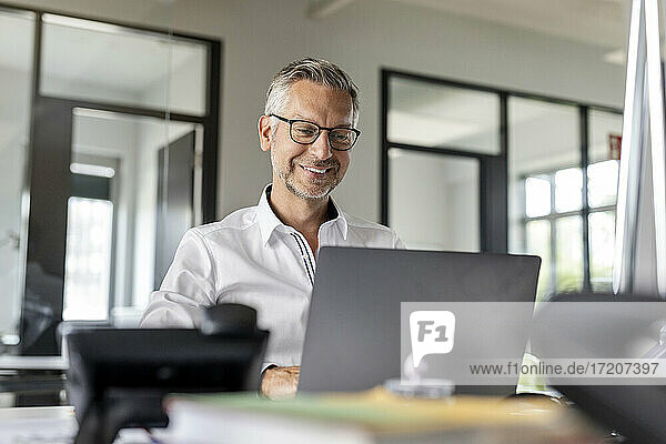 Lächelnder Geschäftsmann  der einen Laptop benutzt  während er am Schreibtisch im Büro sitzt