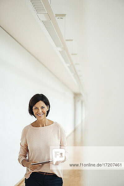 Lächelnde Geschäftsfrau  die ein digitales Tablet auf dem Flur eines Büros hält