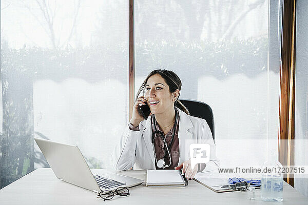 Lächelnde Ärztin  die wegschaut  während sie mit einem Smartphone in einer medizinischen Klinik spricht