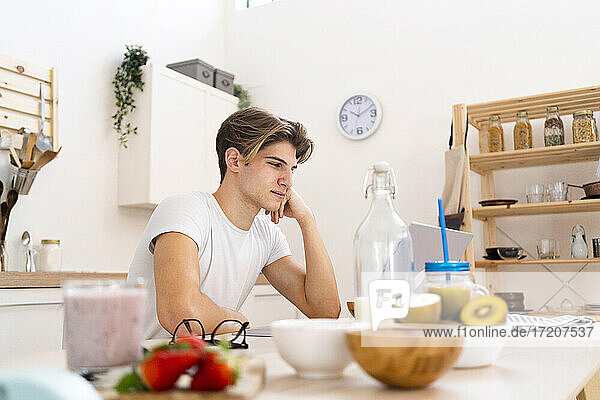 Nachdenklicher junger Mann sitzt mit Laptop in der Küche zu Hause