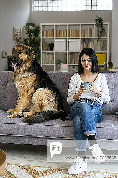 Junge Frau hält Kaffeetasse  während sie mit Hund auf dem Sofa zu Hause sitzt