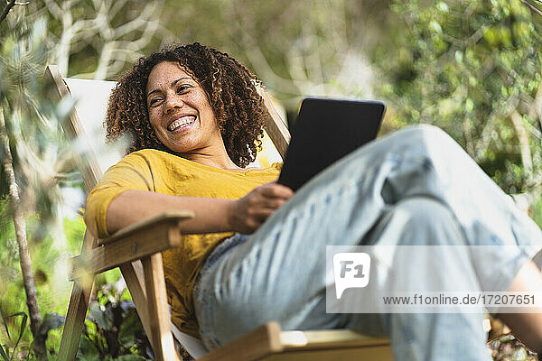 Lächelnde Frau mit digitalem Tablet  die wegschaut  während sie auf einem Liegestuhl im Gemüsegarten sitzt