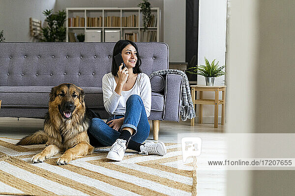 Smiling woman talking on phone while sitting with dog on carpet at home