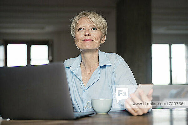 Lächelnder Freiberufler mit Laptop  der wegschaut  während er sein Handy am Schreibtisch im Home Office hält