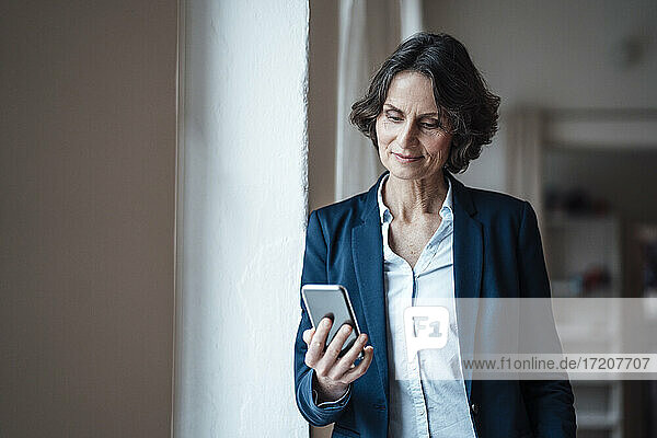 Geschäftsfrau hält Smartphone  während sie sich im Büro zu Hause an die Wand lehnt