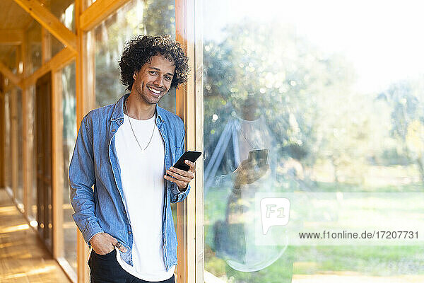 Smiling young man holding smart phone while standing with hand in pocket at front yard
