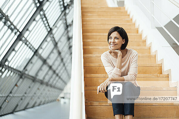 Lächelnde Geschäftsfrau mit Hand am Kinn auf einer Treppe im Korridor sitzend