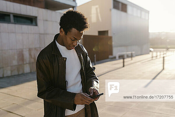 Mittelgroßer erwachsener Mann  der während des Sonnenuntergangs auf dem Fußweg steht und mit seinem Smartphone SMS schreibt
