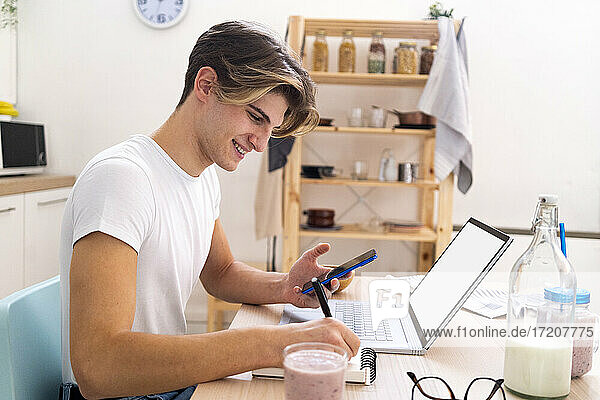 Glücklicher Mann  der mit Laptop und Mobiltelefon in der Küche sitzt und auf einen Notizblock schreibt