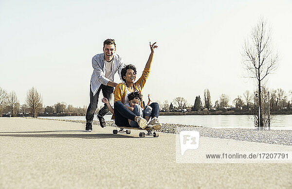 Man pushing woman sitting with boy on skateboard during sunny day