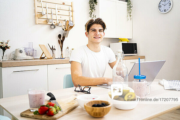 Young man with laptop sitting at table in kitchen