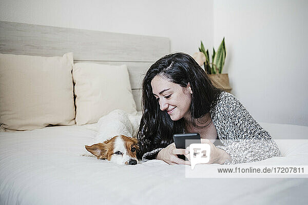 Lächelnde Frau mit Mobiltelefon neben Jack Russell Terrier auf dem Bett liegend