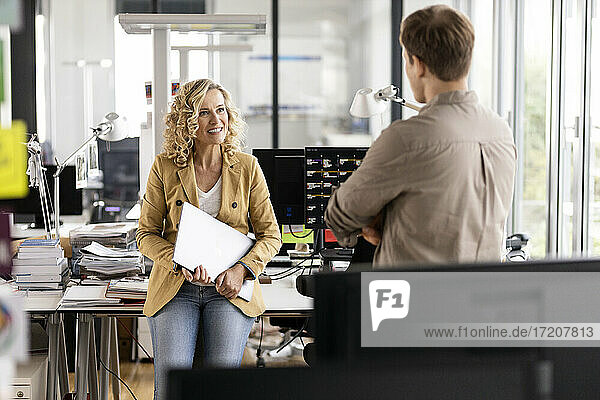 Lächelnde Geschäftsfrau mit Laptop  die mit einem jungen Kollegen diskutiert  während sie sich an einen Schreibtisch im Büro lehnt